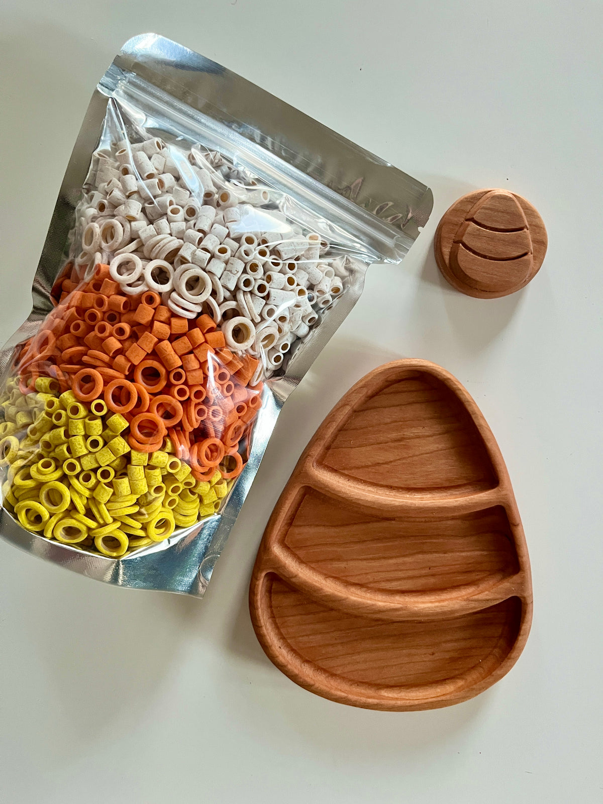 Silver resealable bag with small colorful clay beads next to a three-compartment wooden tray and round wooden lid on white surface