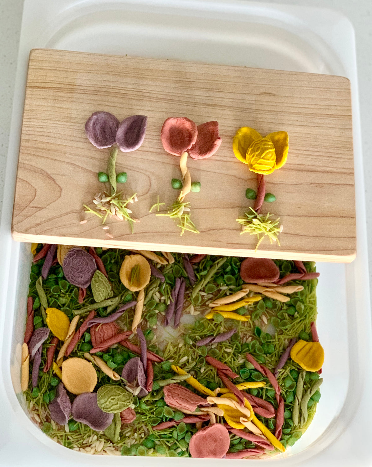 Multicolored flower shapes made from pasta and peas arranged on a wooden board above matching pasta pieces in a sensory bin