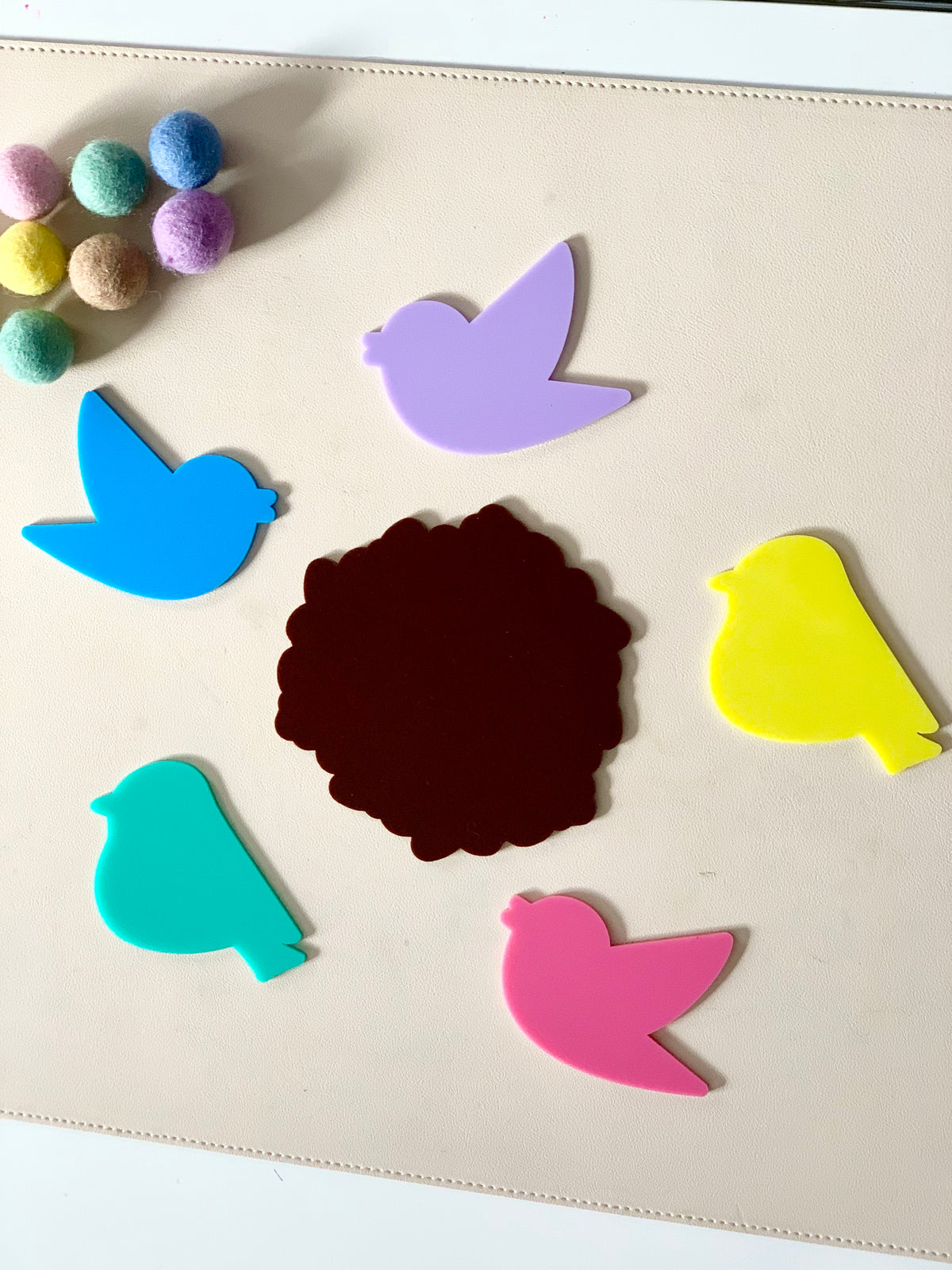Colorful bird-shaped cutouts arranged around a dark brown scalloped circle with pastel pom-poms nearby on a light surface