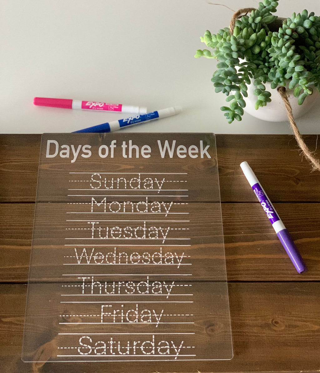 Clear acrylic tracing board with dotted days of the week and three colored dry erase markers on wooden table next to plant