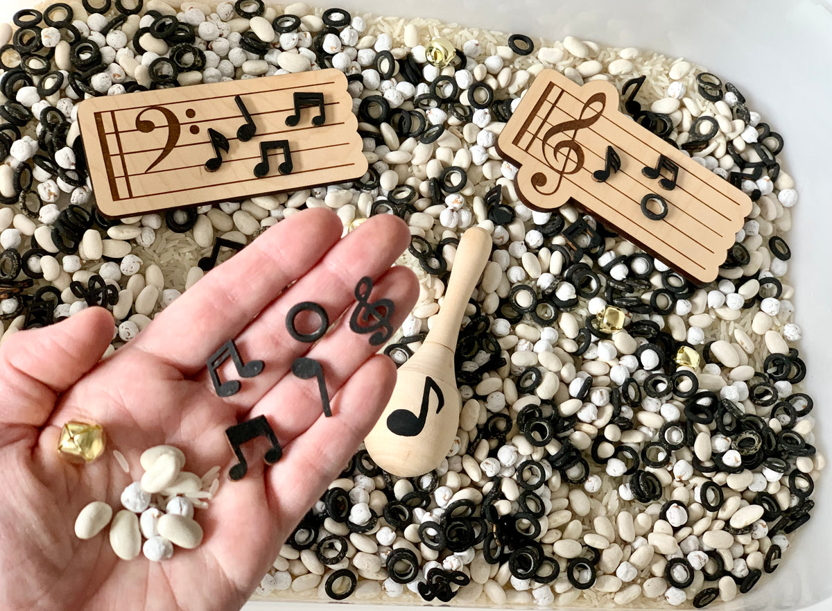 Hand holding small black musical note tokens above a sensory bin filled with white and black beans and wooden music staff plaques and maraca