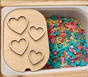 Wooden sensory tray with four heart-shaped cutouts next to bin filled with colorful pasta and dried corn kernels