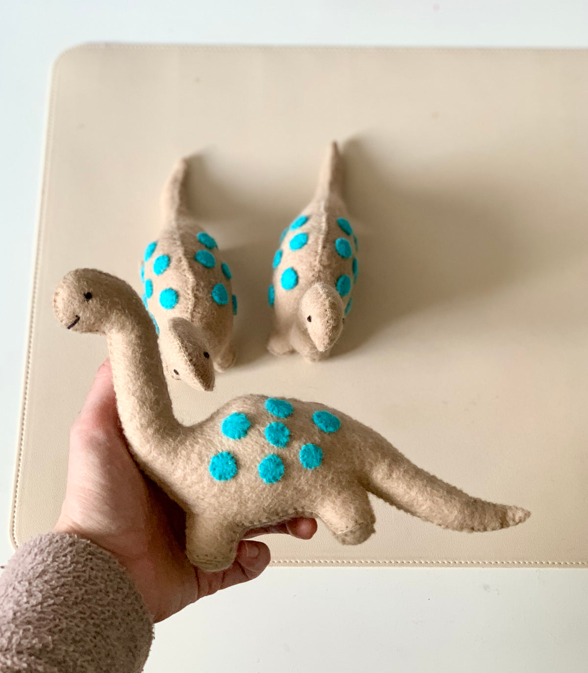 Beige felt dinosaur toy with blue felt spots held in hand with two similar toys on a cream surface in the background