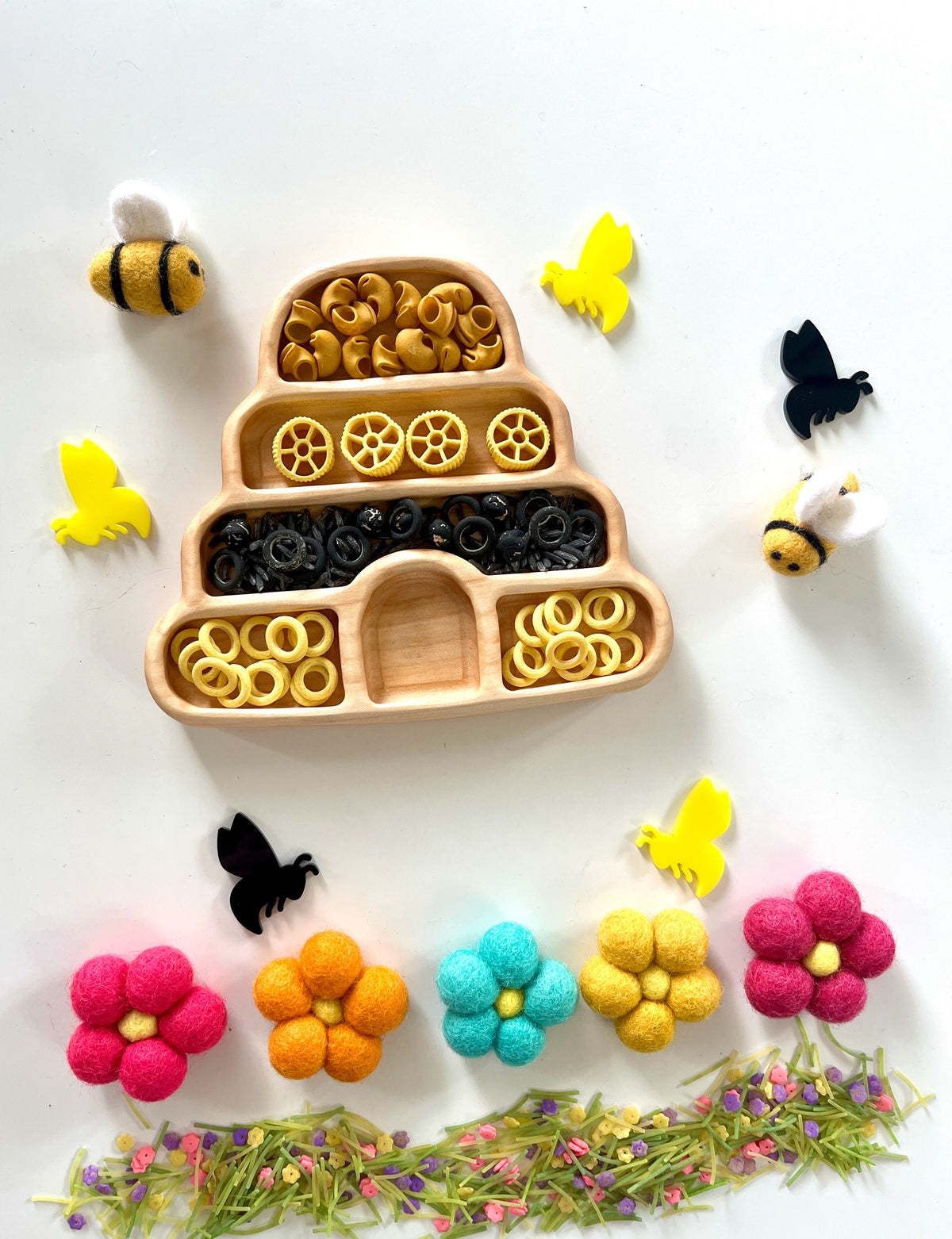 Wooden beehive tray filled with yellow and black pasta, surrounded by felt bees, flowers, and yellow and black bee cutouts on white background