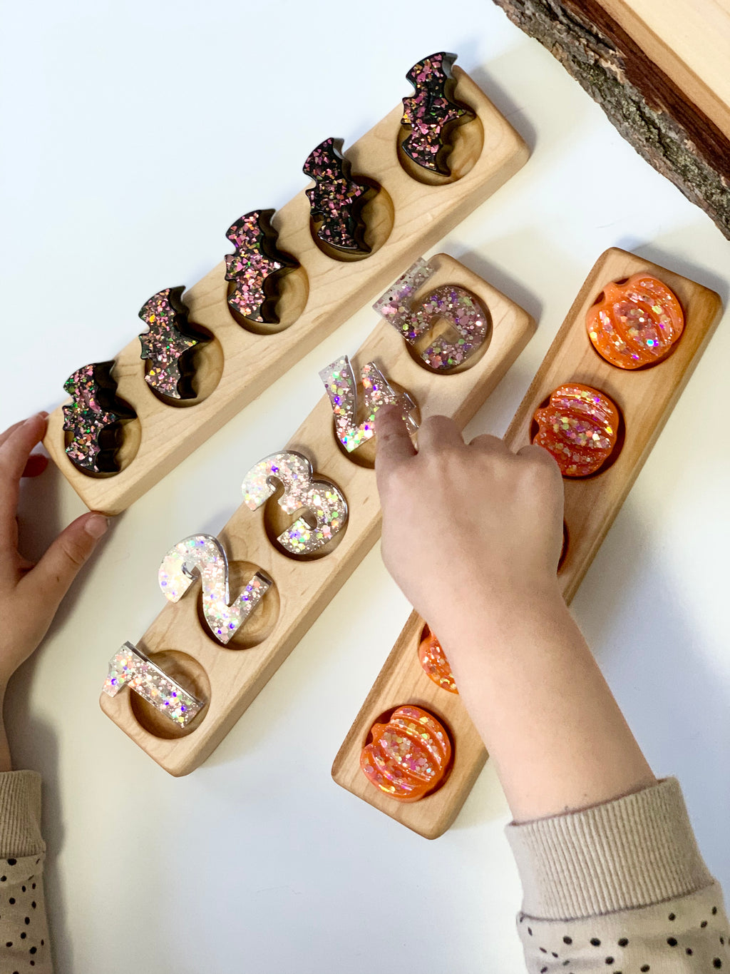 Three wooden five frame sensory trays with glittery bat, pumpkin, and number pieces held by child’s hands on white surface