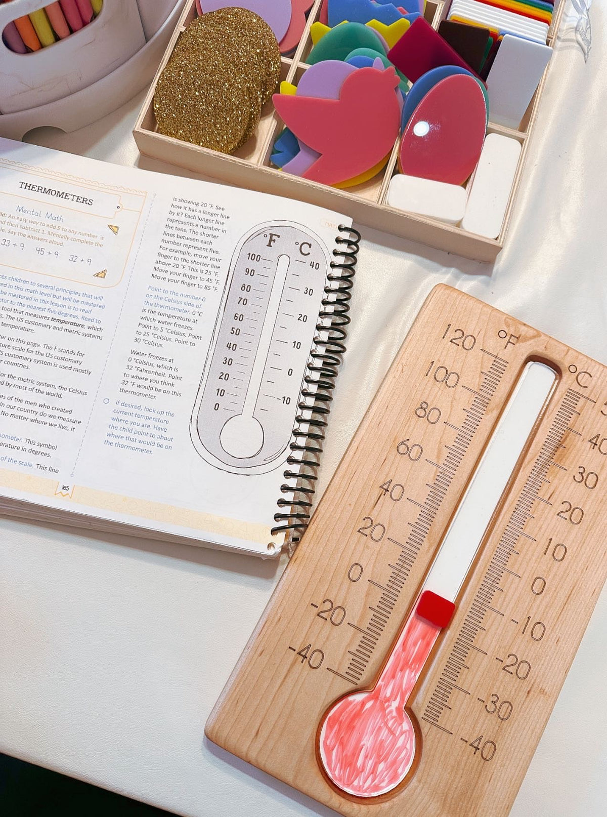 Wooden thermometer with red liquid indicator next to an open educational book and colorful foam shapes in compartments