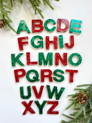 Red and green glittery resin alphabet letters arranged on white background with pine branches and cones nearby