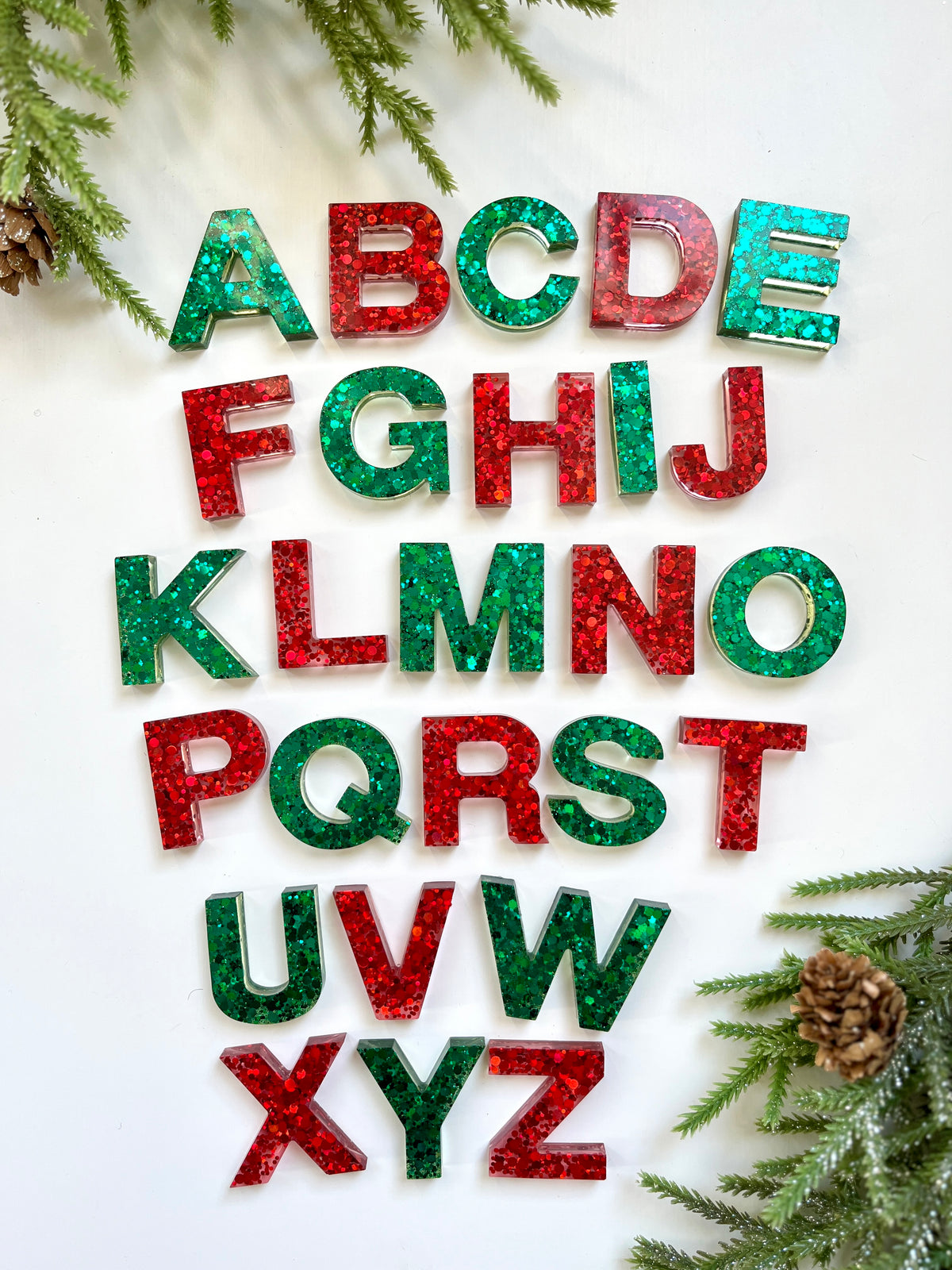 Red and green glittery resin alphabet letters arranged on white background with pine branches and cones nearby