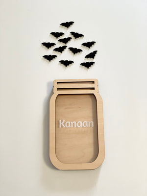 Light wooden jar-shaped frame with the name Kanaan and black bat cutouts arranged above on a white background