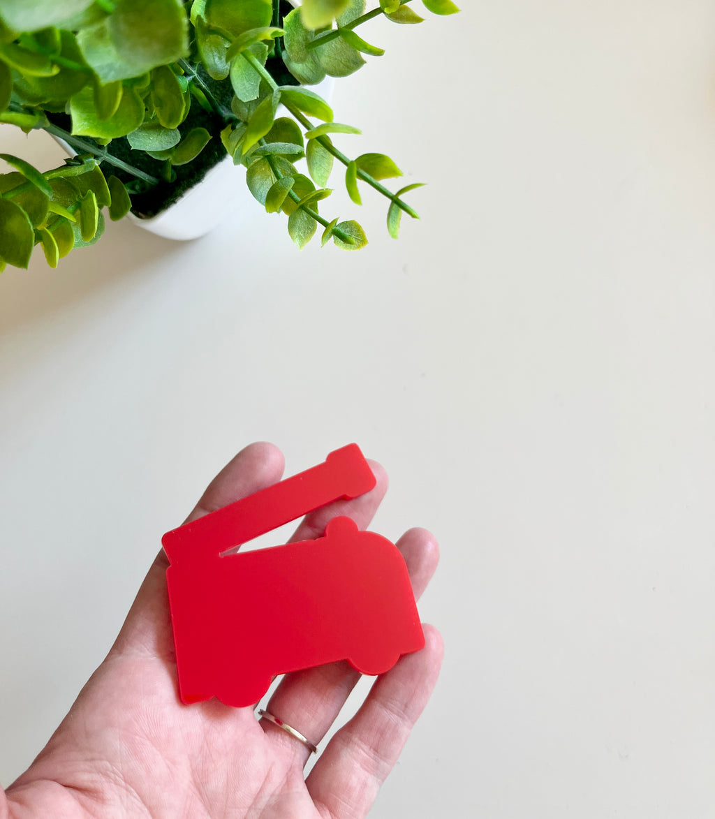 Hand holding a smooth red fire truck-shaped cutout with green leaves from a potted plant above on a white surface