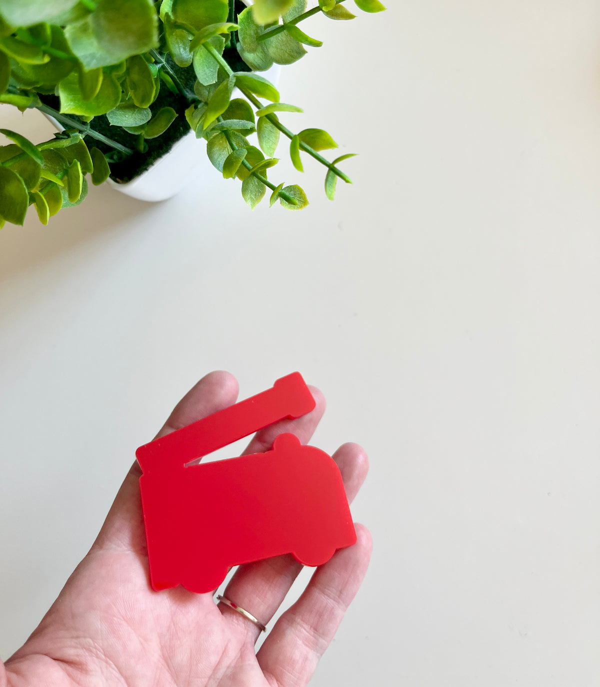 Hand holding a smooth red fire truck-shaped cutout with green leaves from a potted plant above on a white surface