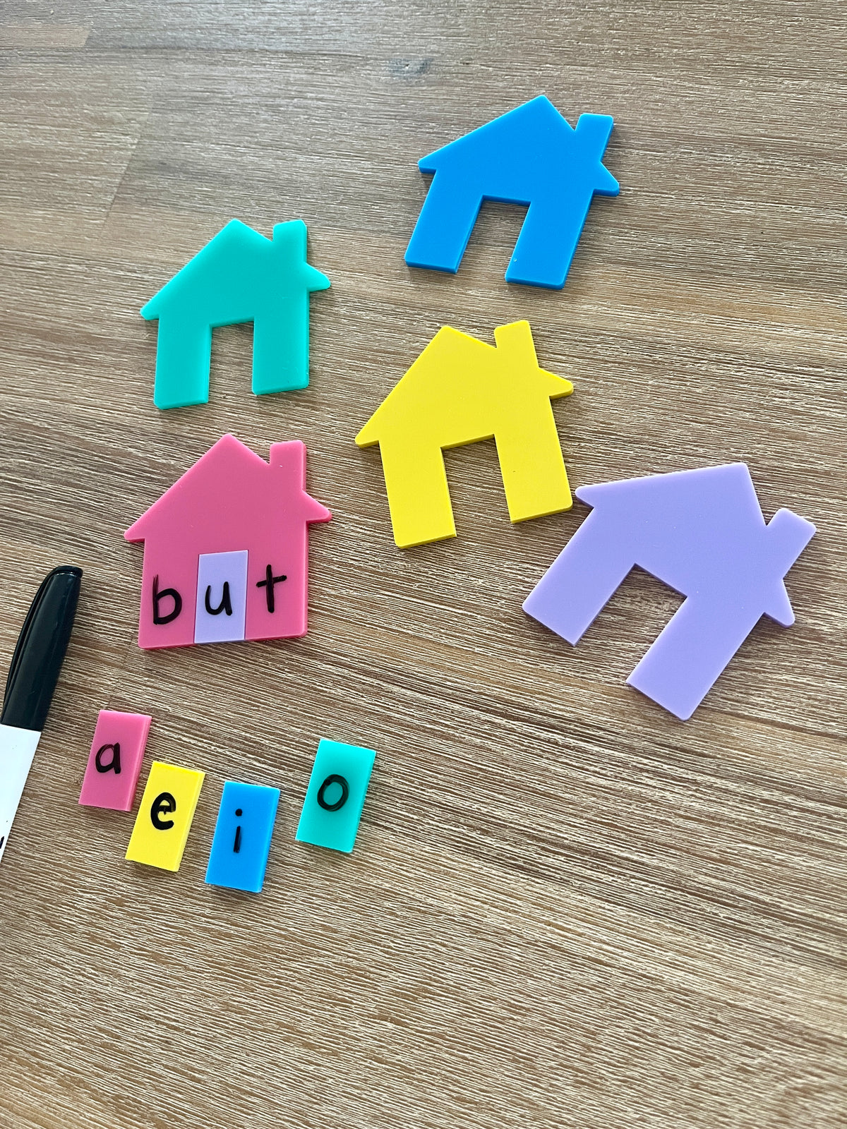 Little Dry Erasables - Houses for CVC, Math + More featuring colorful plastic house shapes and letter tiles spelling "but" with vowel tiles and a black marker on a wood surface, handcrafted homeschool education resources.