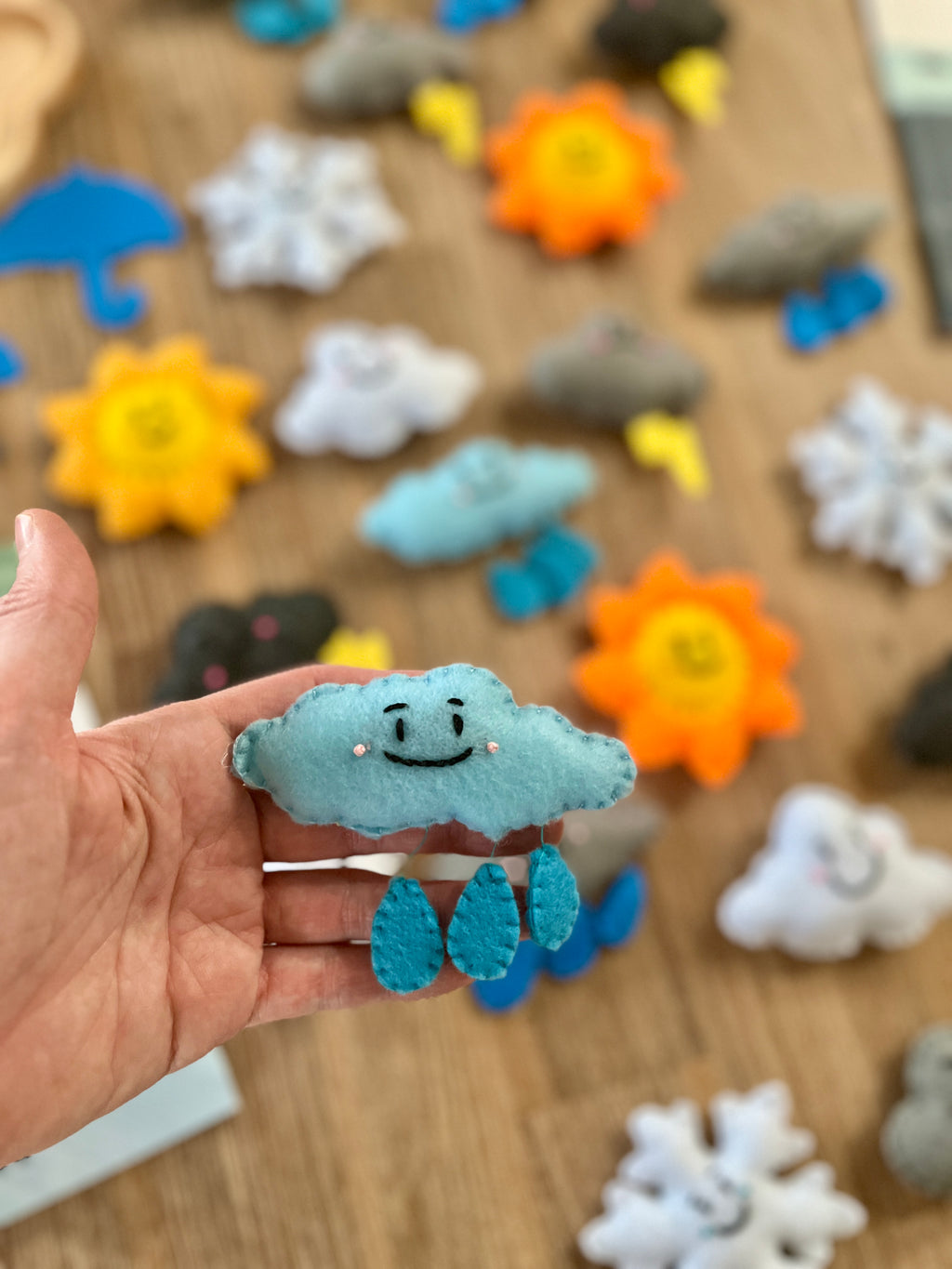 Hand holding a smiling blue felt cloud with raindrop details, with a background of various colorful Felt Weather Shapes, Made in Canada, handcrafted, homeschool, education resources.