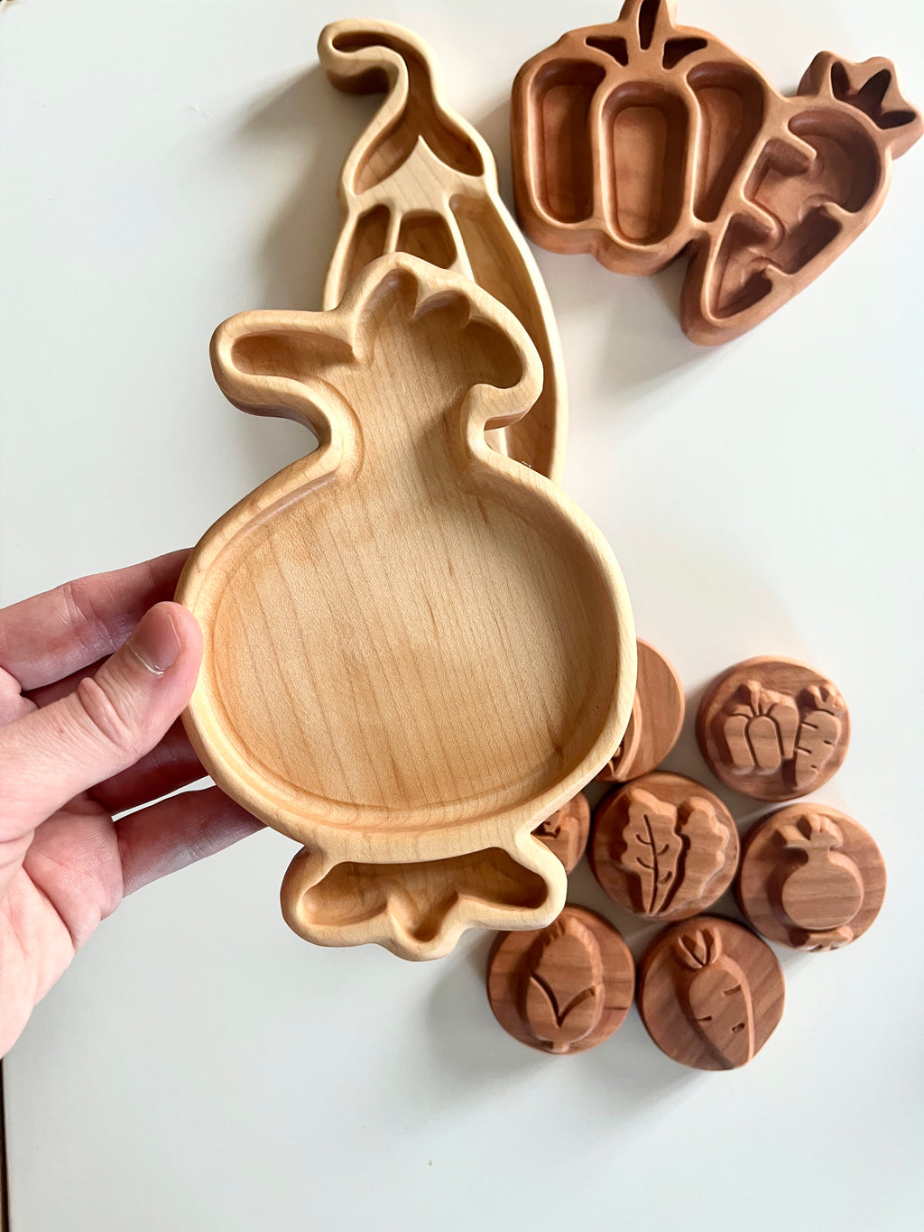 Hand holding wooden tray shaped like a pomegranate next to wooden trays and round tokens with vegetable carvings on white surface