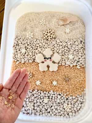 Winter sensory kit with layered rice, beans, wooden and felt snowflakes, and gold flakes in hand in plastic tray