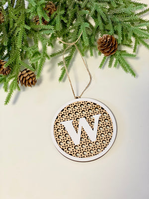 Round wooden Christmas ornament with monogram letter W and rattan design hanging near pinecones and green branches