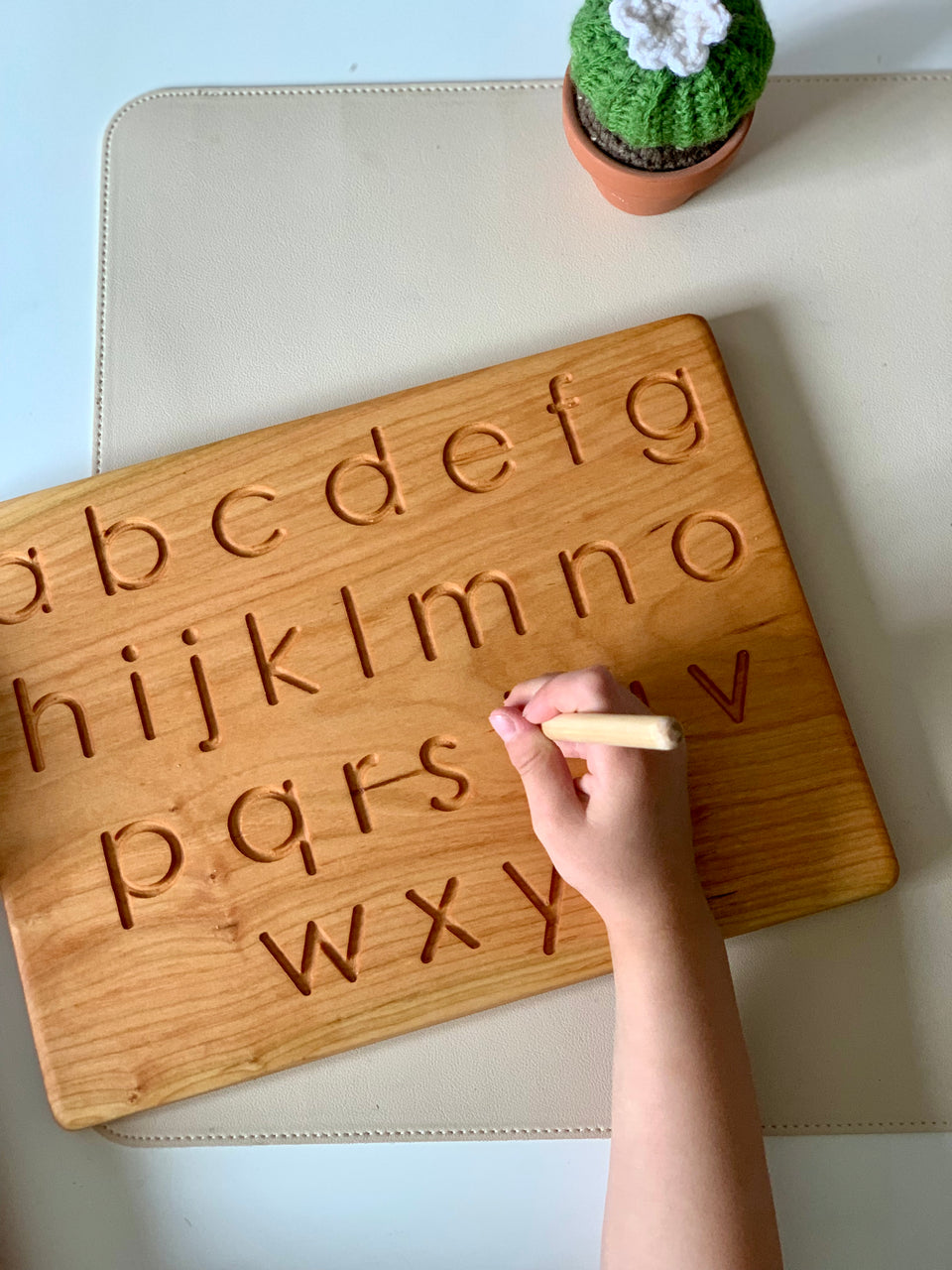 Child’s hand tracing carved lowercase alphabet letters on wooden board with a small green felt cactus nearby