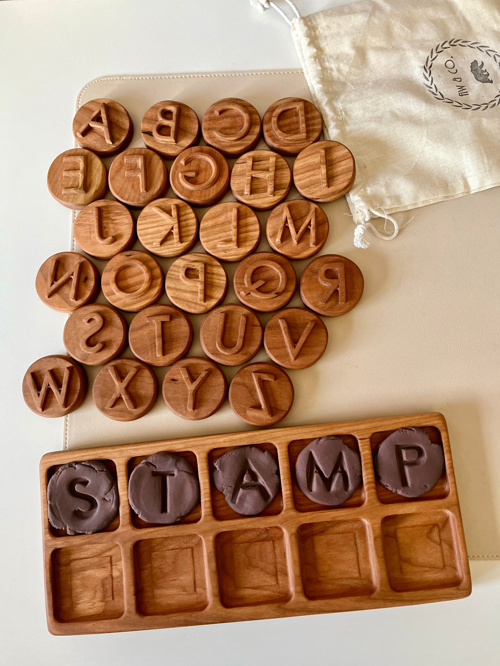 Round wooden alphabet discs arranged beside a wooden tray holding letter-shaped brown clay stamps and a cloth drawstring bag