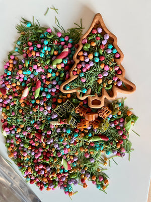 Wooden tree-shaped tray filled with colorful beads, dried green pine needles, and spiral striped pasta on white background