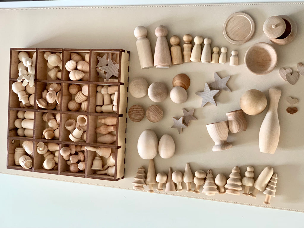 Variety of smooth natural unfinished wooden shapes including peg dolls, stars, trees, bowls, and spheres on light surface