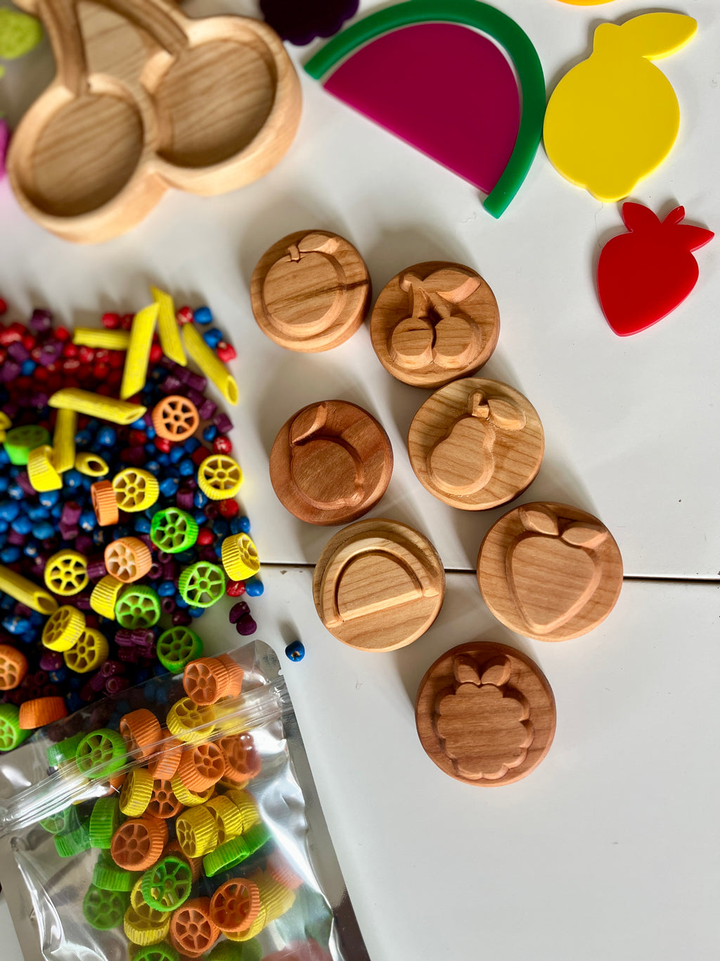 Wooden discs carved with fruit shapes scattered on white surface near colorful pasta pieces and plastic fruit cutouts