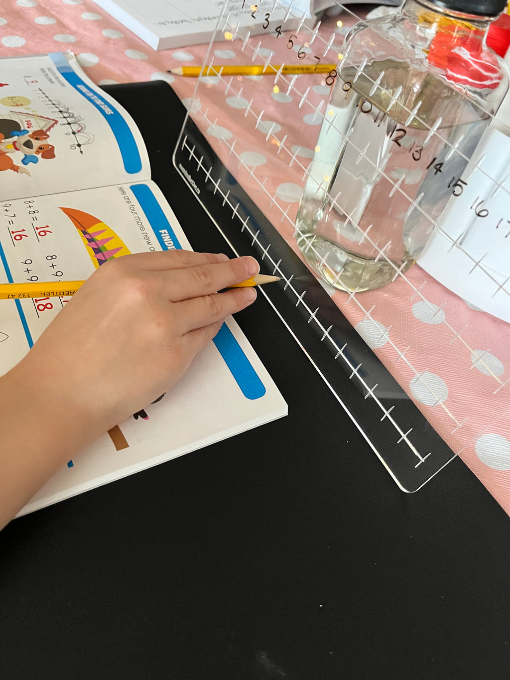 Child writing on blackboard beneath clear acrylic number line numbered 0 to 16 on pink polka dot surface with bottle nearby