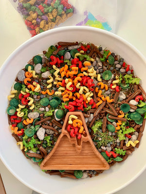 Wooden volcano dish with red, orange, yellow curly pasta, green leaves, and gray pebble-shaped play pieces in white bowl