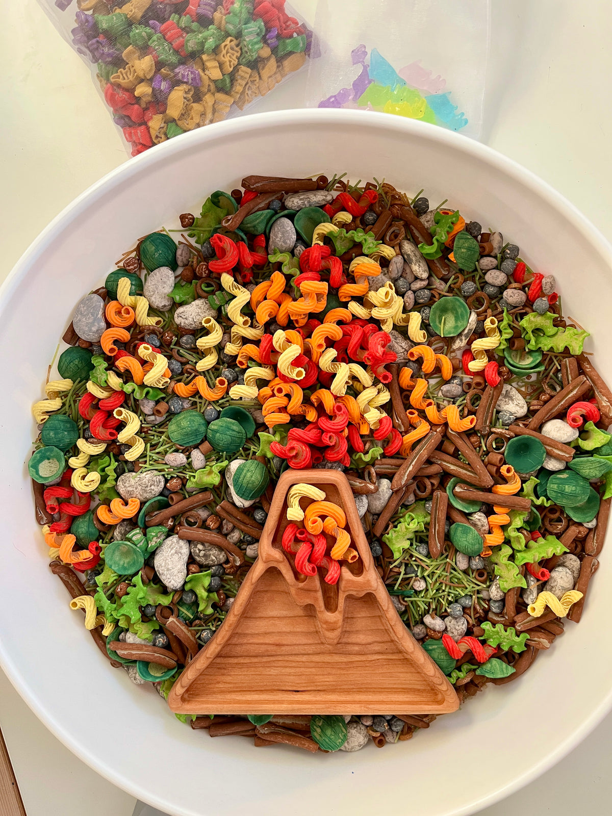 Wooden volcano dish with red, orange, yellow curly pasta, green leaves, and gray pebble-shaped play pieces in white bowl