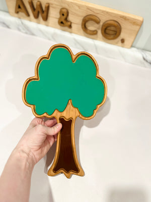 Hand holding a wooden tray shaped like a tree with green leafy top and brown trunk on a white surface