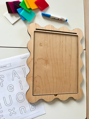 Wooden-framed dry erase board with scalloped edges, colorful translucent squares, marker, and alphabet practice sheet nearby