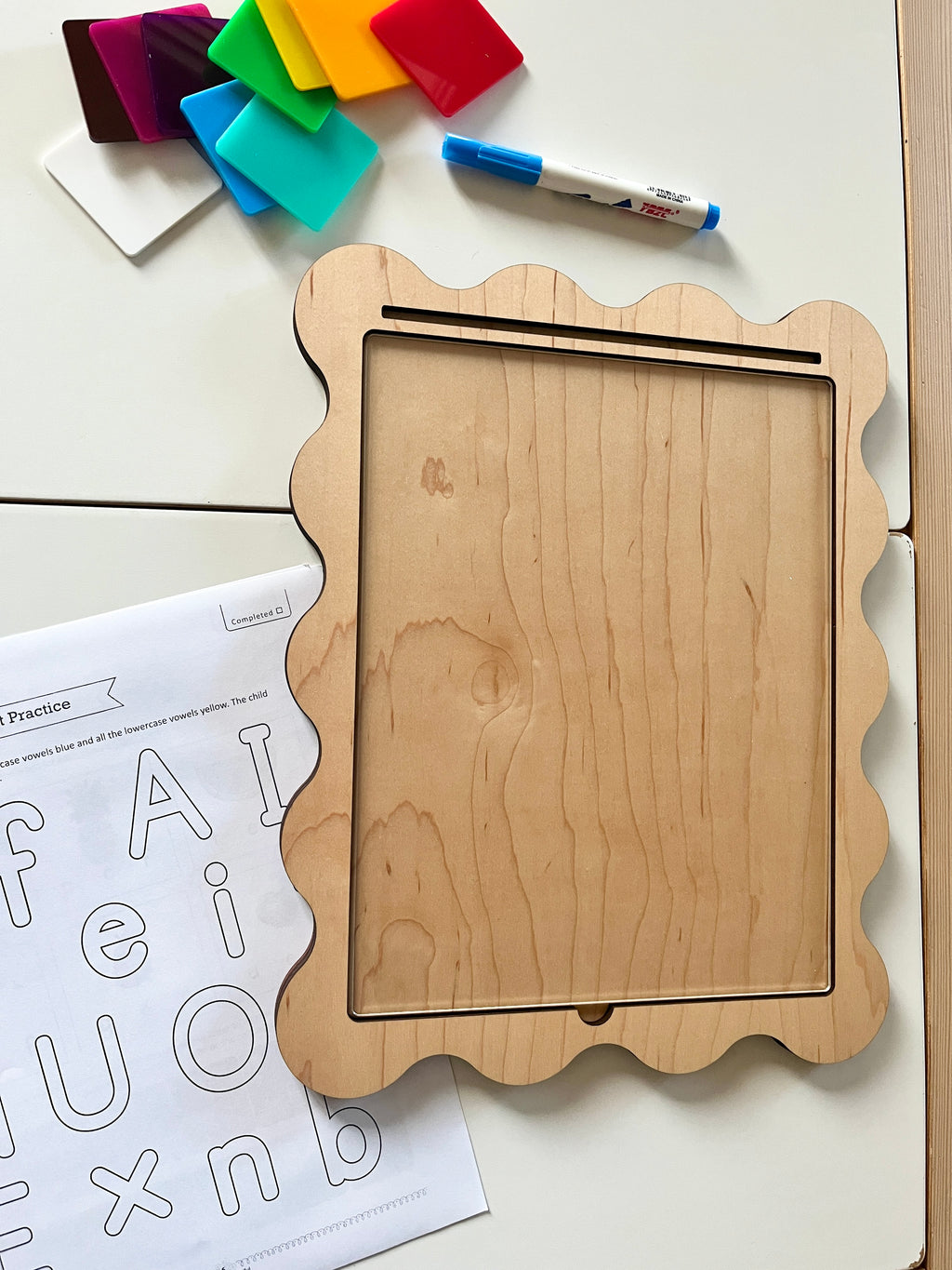 Wooden-framed dry erase board with scalloped edges, colorful translucent squares, marker, and alphabet practice sheet nearby