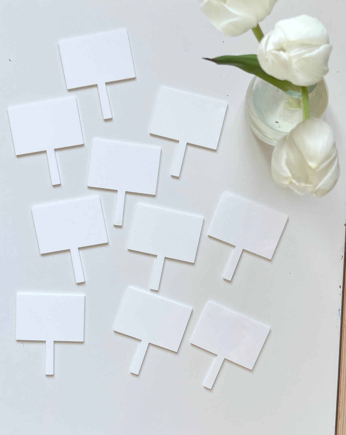 Ten blank white rectangular plant labels spread on a white surface near a glass vase with white tulips
