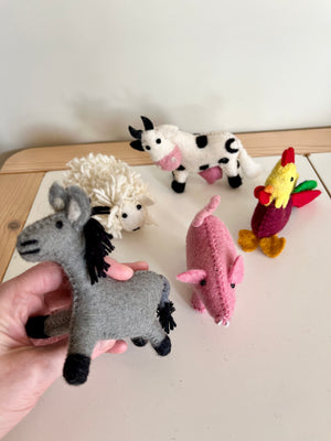 Five handmade felt farm animal toys including a gray donkey held in hand with sheep, cow, pig, and rooster on a white surface