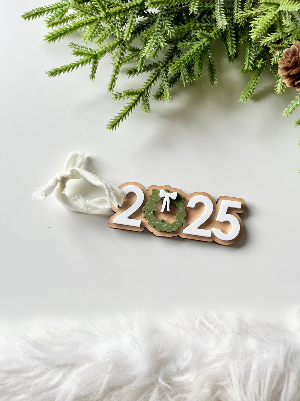 Decorative wooden 2025 ornament with green wreath and white bow tied with white fabric ribbon on white surface