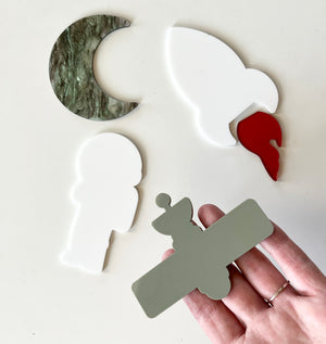 Four acrylic shapes including a green marble crescent moon, white bird with red beak, white astronaut, and gray satellite held on fingers