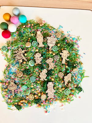 Wooden nutcracker and ballerina figurines on green pine needle mix with pastel sprinkles and gold beads on white surface