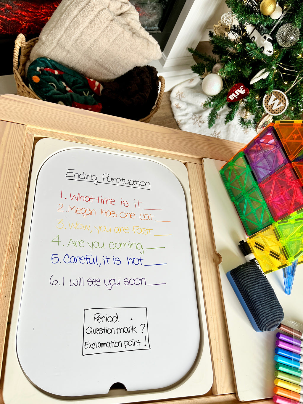 Whiteboard with colorful handwriting showing sentences for punctuation practice beside translucent magnetic tiles and markers on wood tray