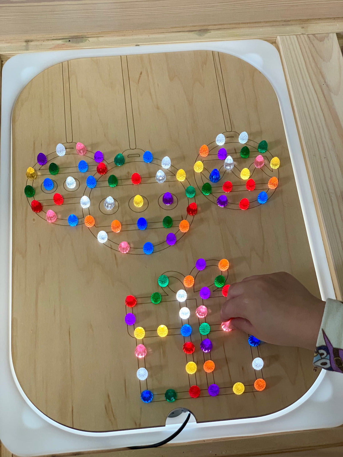 Wooden Flisat insert with Christmas ball and gift shapes lit up by colorful light pegs with child’s hand placing a peg