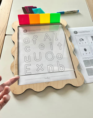 Wooden-framed dry erase board with colorful magnets and alphabet practice worksheet underneath clear acrylic sheet