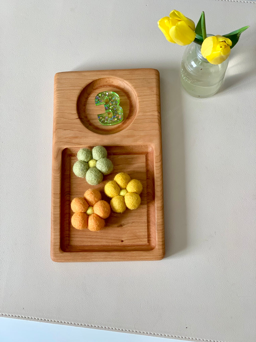 Wooden tray with glittery number three and three felt flowers in green, orange, and yellow next to a small jar with yellow tulips