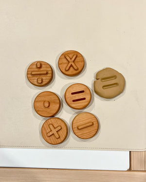 Seven round wooden tokens with mathematical symbols and a matching clay piece with an equal sign imprint on a beige surface