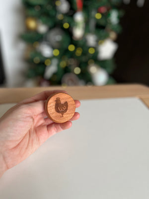Hand holding round wooden token with engraved chicken design against blurred holiday tree background