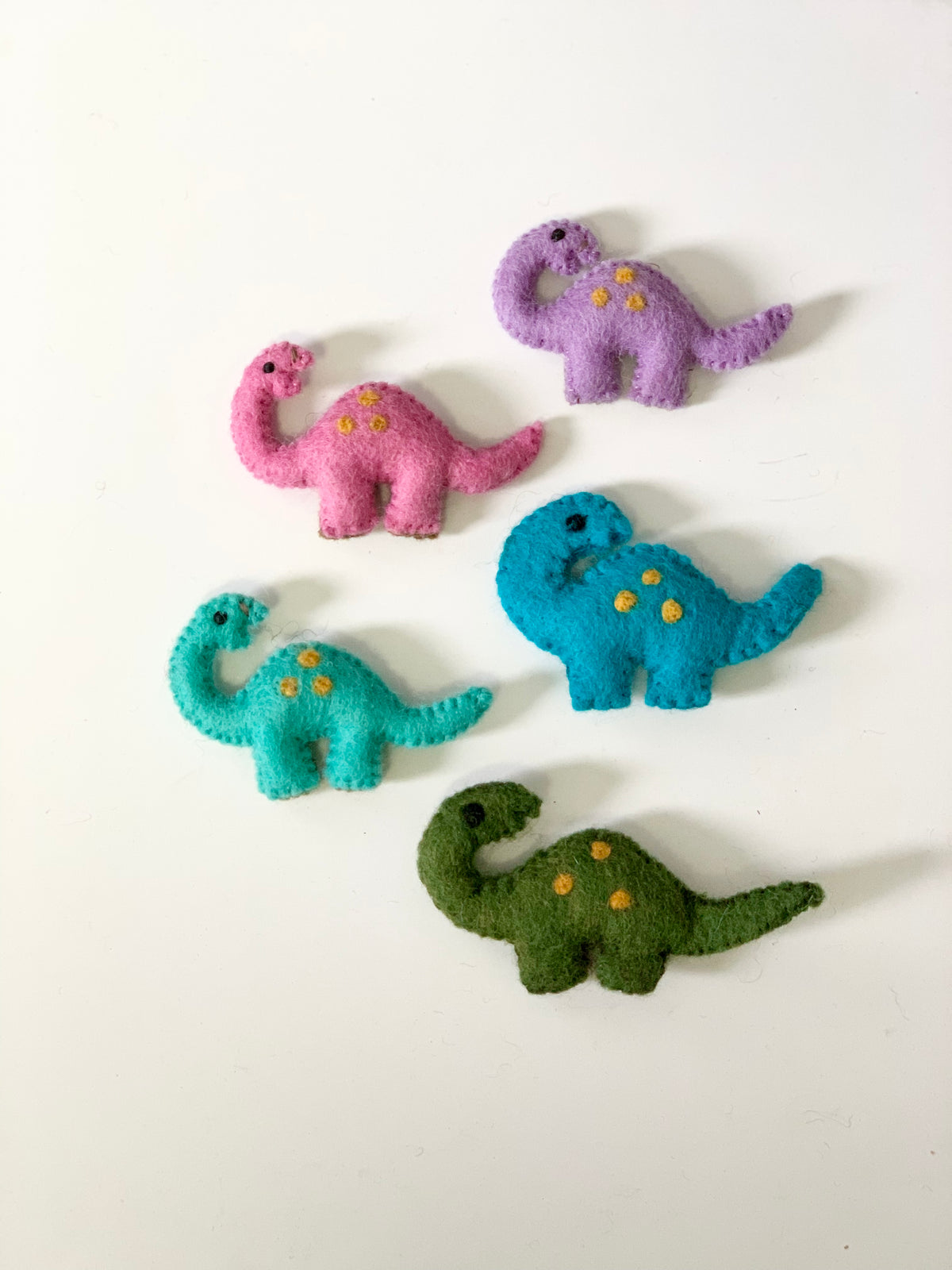 Set of five handmade felt dinosaurs in purple, pink, blue, teal, and green with yellow spots on white background