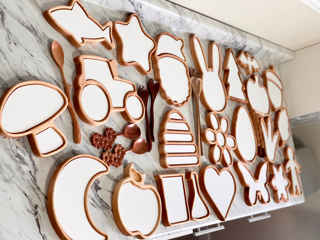 Wooden sensory trays shaped like animals, flowers, fruit, and objects with white inserts on marble surface