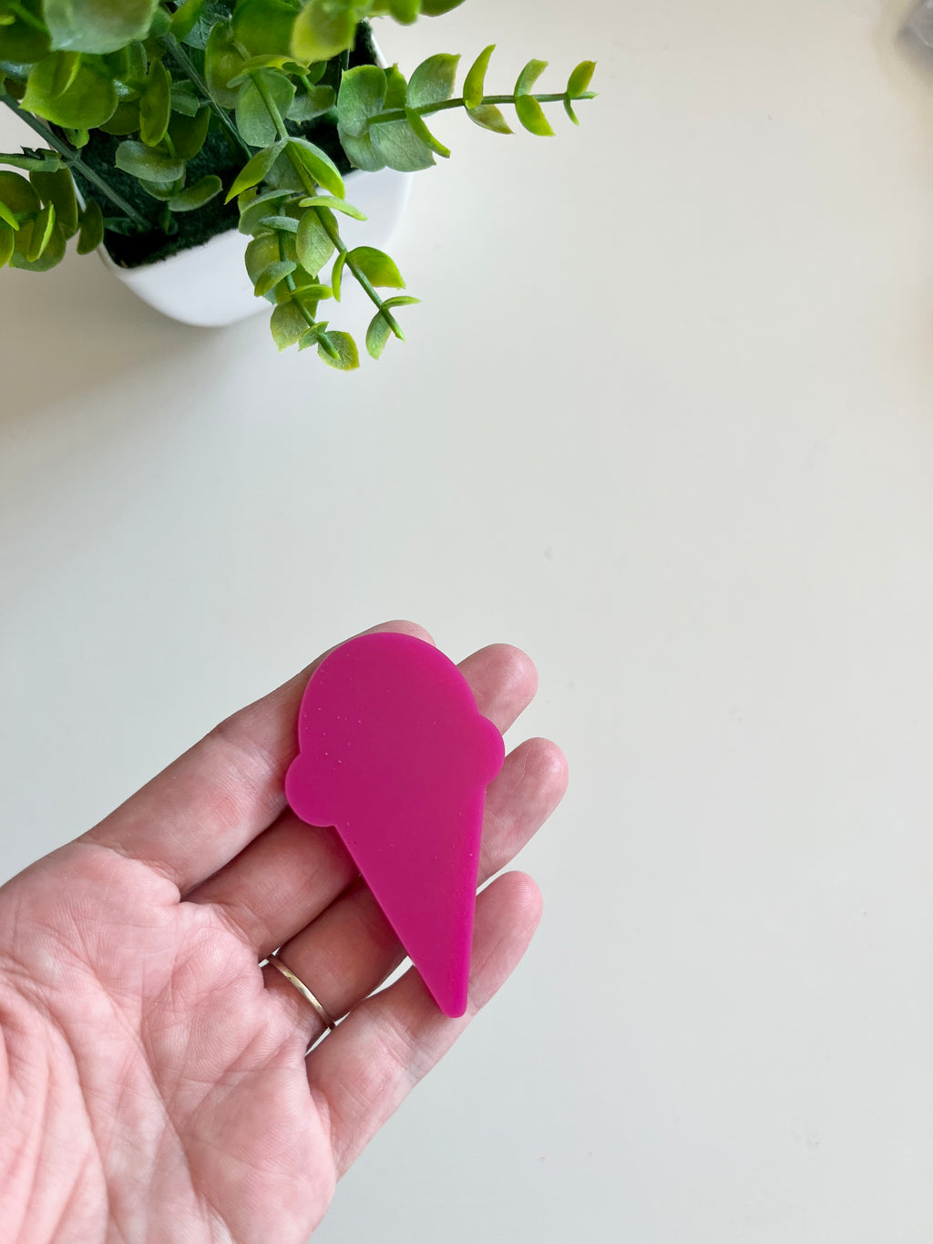 Hand holding a smooth magenta ice cream cone-shaped cutout with a green potted plant in the background on a light surface