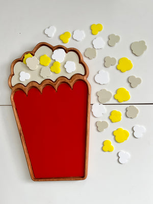 Wooden tray shaped like popcorn container with red base and scattered yellow, white, and beige popcorn-shaped tokens on white surface