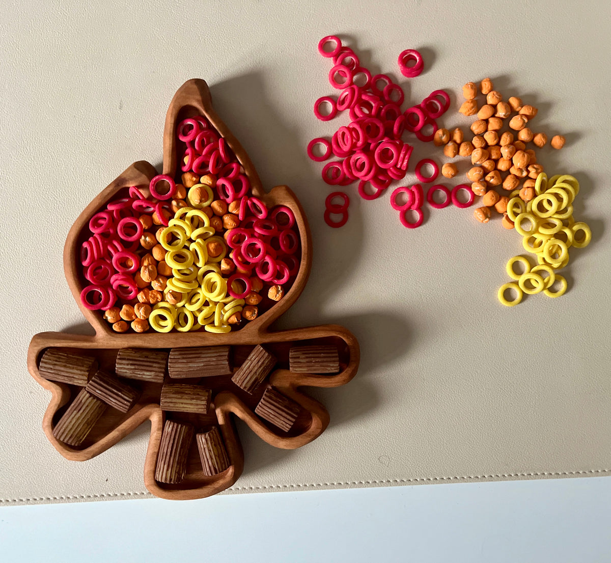 Wooden campfire-shaped tray filled with red, yellow, and orange ring and bead-shaped pieces on a beige surface