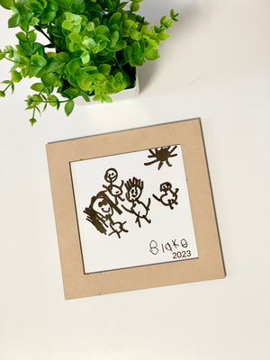 Framed laser-cut square featuring a child’s drawing traced by hand with figures and sun on white background