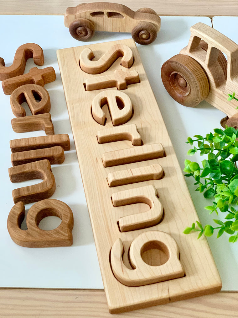 Personalized hardwood name puzzle with lowercase wooden letters spelling Sterling on natural wood board with wooden vehicles and green plant