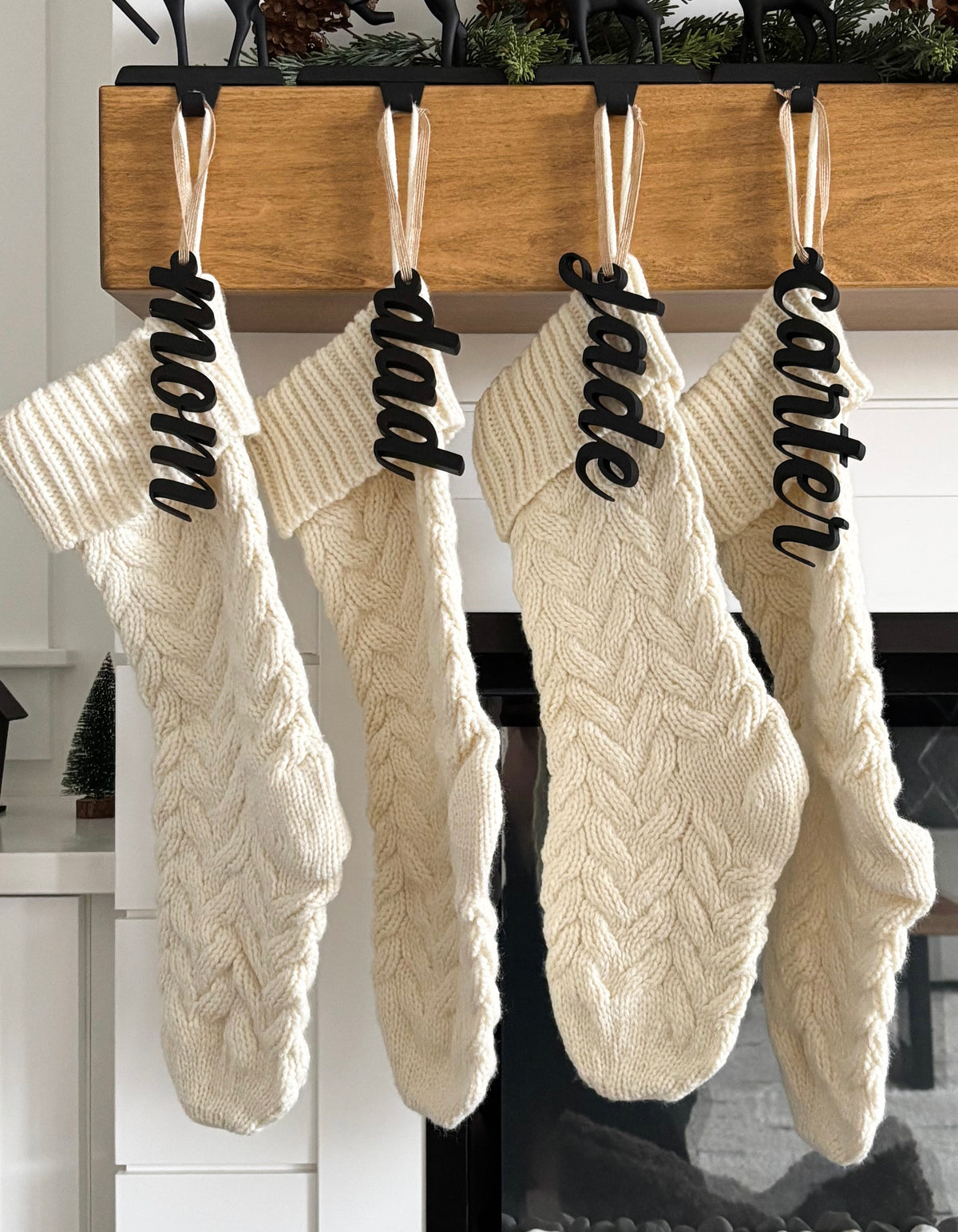 Four cream-colored cable knit Christmas stockings hanging from a wooden mantel with black name tags and white ribbons
